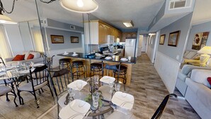 A modern kitchen with wooden cabinets, a central island, and bar stools. There is a dining area with a glass table and chairs. - SUNRISE /SUNSET GULF VIEWS #932 - FREE BEACH CHAIRS!!!!! (Panama City)