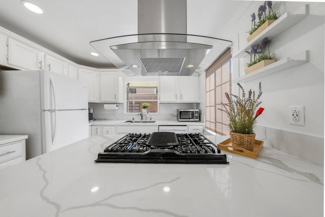 Une cuisine moderne avec des meubles blancs, une hotte en acier inoxydable, un îlot central doté d’une cuisinière à gaz, d’un four à micro-ondes et d’une plante en pot.
