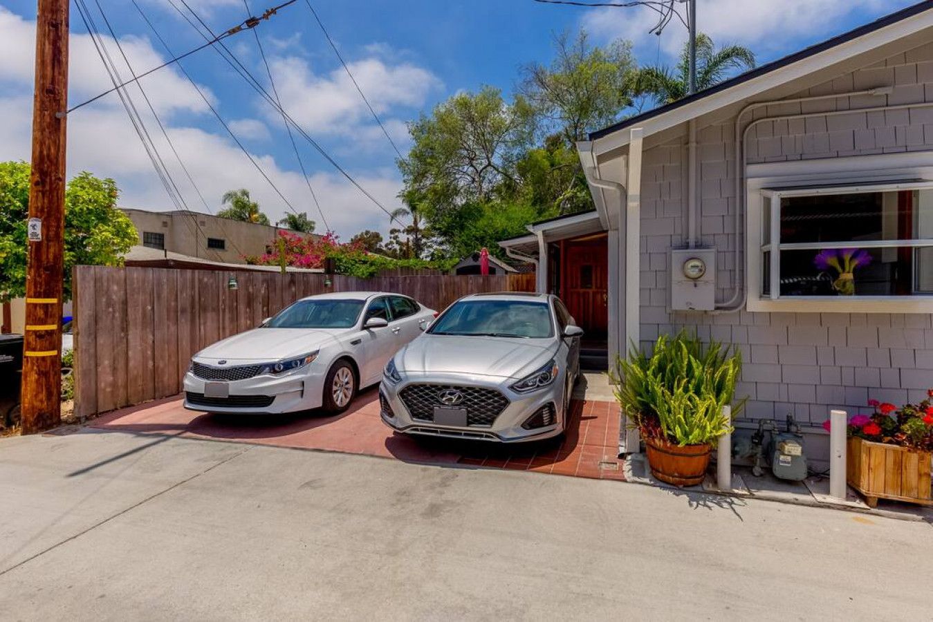 A residential driveway with two cars parked, a wooden fence, and a house with a window and a potted plant.