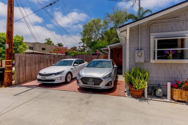 A residential driveway with two cars parked, a wooden fence, and a house with a window and a potted plant.