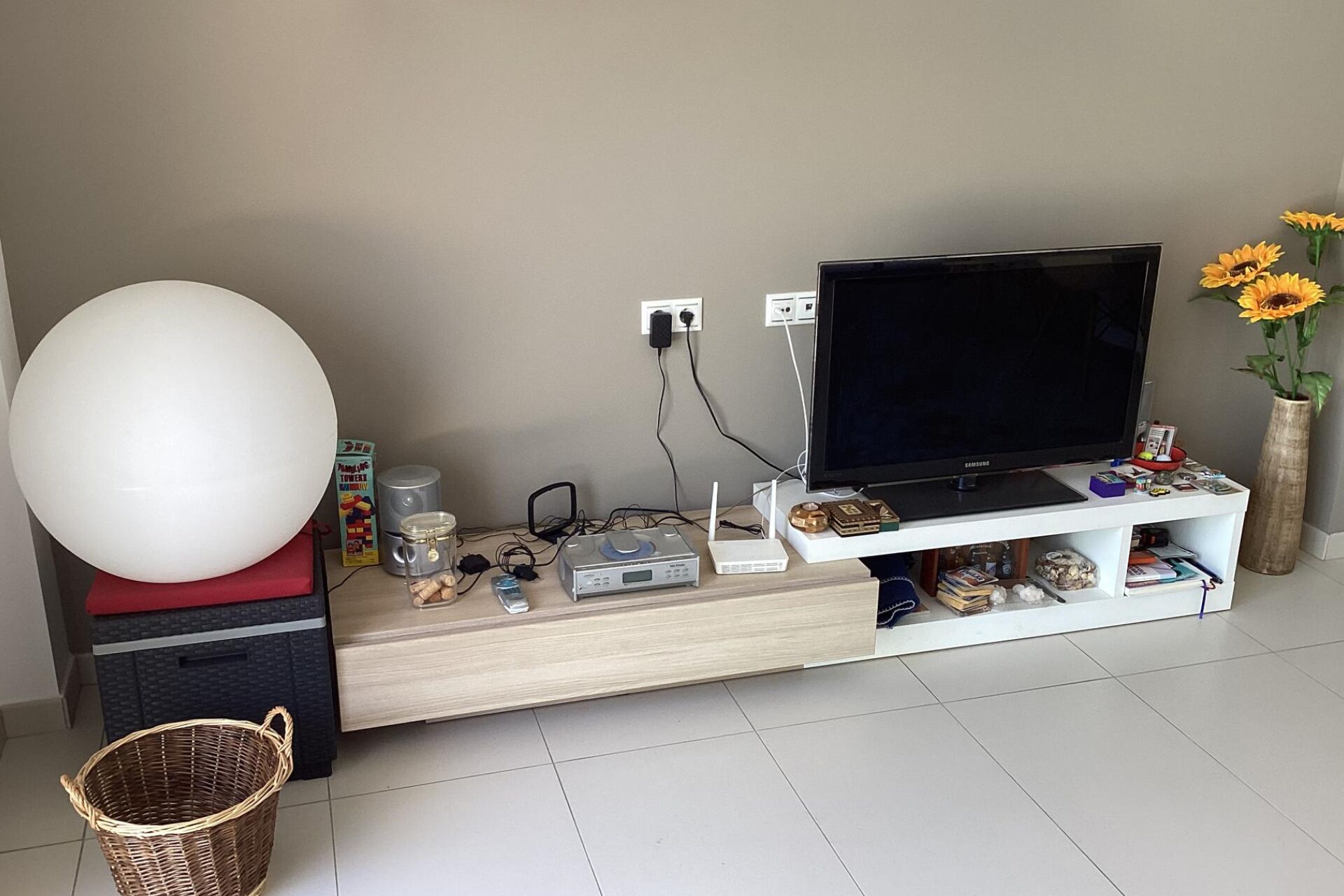 Een moderne woonkamer met een flatscreen televisie op een witte tv-meubel, een grote witte bolvormige lamp en een vaas met zonnebloemen.