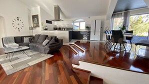 A modern living room with a grey sofa, a coffee table, and a dining area with a table and chairs. A kitchen with white cabinets and a stove is visible in the background.