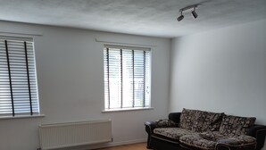 A spacious living room with a wooden floor, a black sofa with patterned cushions, and two windows with blinds. - Immaculate 1-Bed Apartment in Borehamwood (Borehamwood)