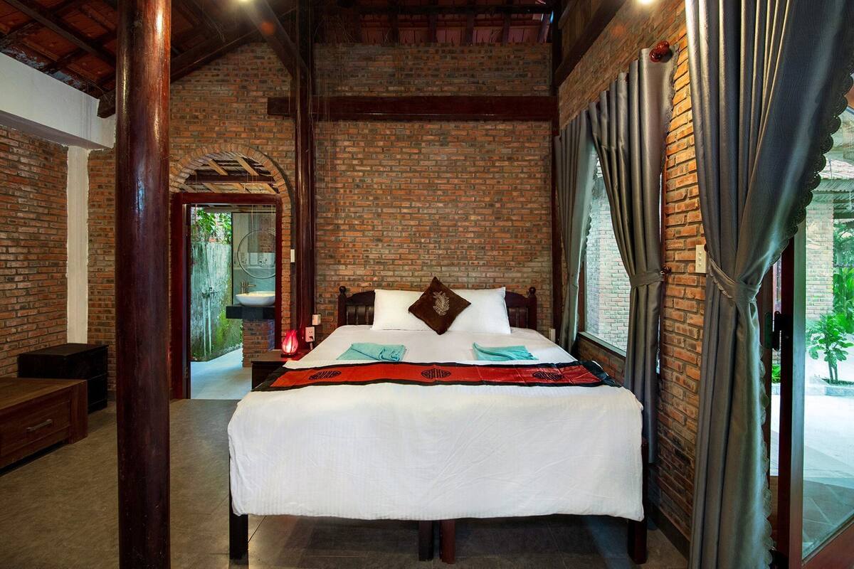 A hotel room with a brick wall, a bed with a white bedspread, and a view of the outdoors.
