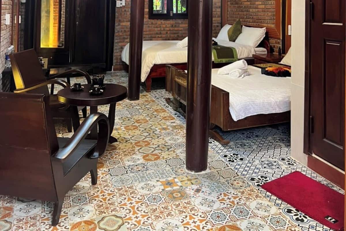 A cozy room with a wooden ceiling, a brick wall, and a tiled floor. There is a bed with pillows and a table with chairs.
