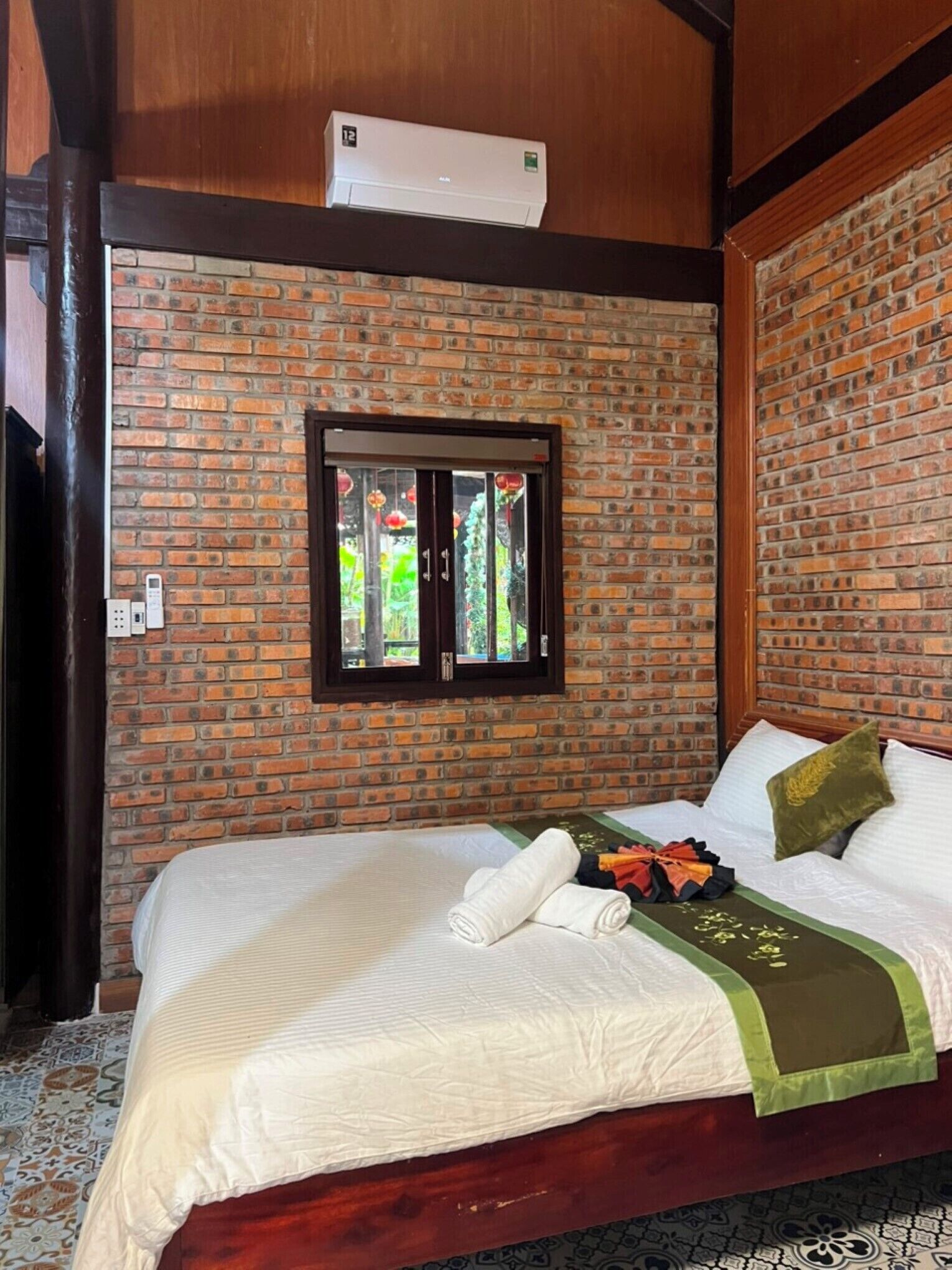 A hotel room with a brick wall, a window with a view of greenery, a bed with white linens, and an air conditioning unit above the window.