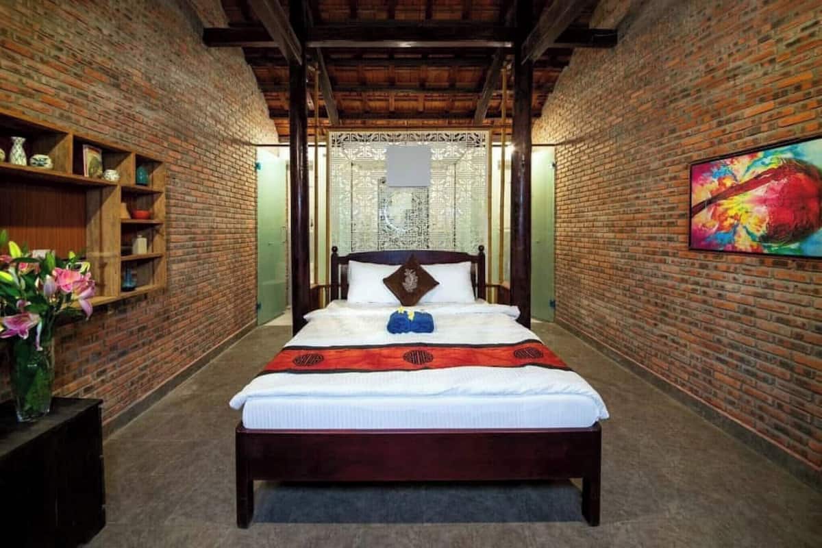 A room with a brick wall, a bed with a wooden frame, a red and black patterned bedspread, and a colorful painting on the wall.