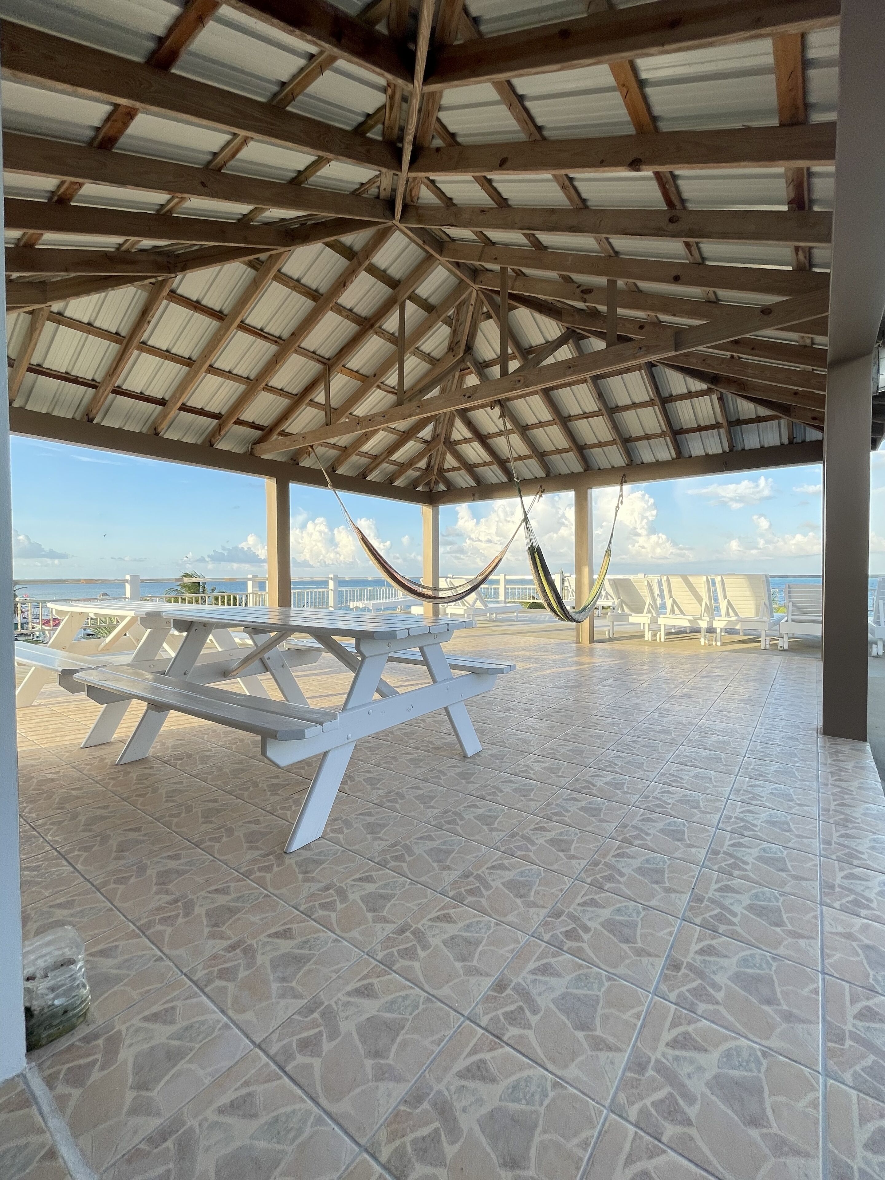 A covered outdoor area with picnic tables and hammocks, overlooking a beach.