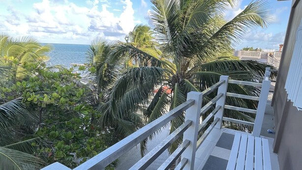 A balcony with a white railing, a white bench, and a view of the sea and palm trees. - Caye Caulker Beach Hotel (Caye Caulker)