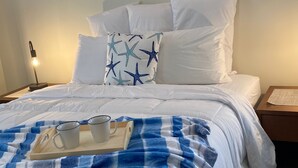 A neatly made bed with white bedding, a blue and white checkered blanket, and a tray with two mugs. There are star-patterned pillows and two seahorse artwork pieces on the wall.