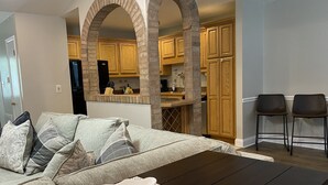 A modern living room with a sofa, a coffee table, and two chairs. There is a kitchen area with wooden cabinets and a brick archway.