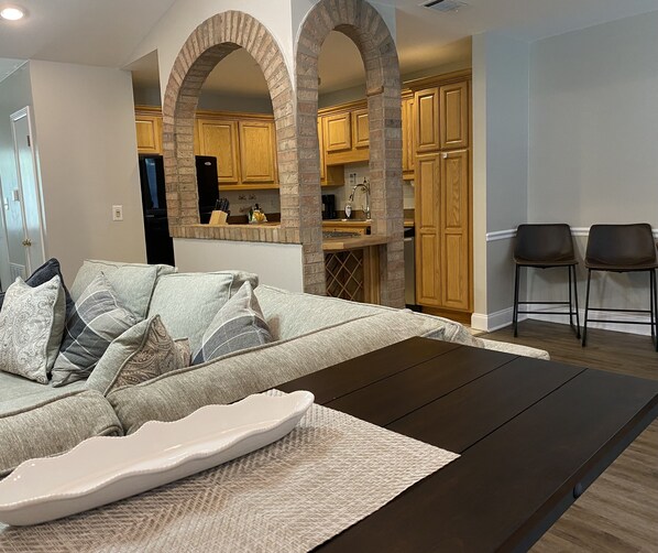 A modern living room with a sofa, a coffee table, and two chairs. There is a kitchen area with wooden cabinets and a brick archway. - Family Retreat w yard, mins to Beach, Convention Center, AF Base. NO CLEAN FEE ! (Fort Walton Beach)