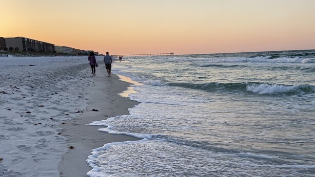 A beach with footprints leading to the water, a pier in the background, and a sunset sky.