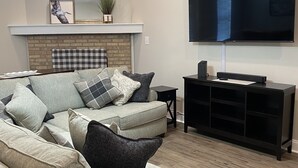 A living room with a grey sofa, a wooden coffee table, a black entertainment unit, a flat-screen TV, a ceiling fan, and a fireplace with a checkered blanket.