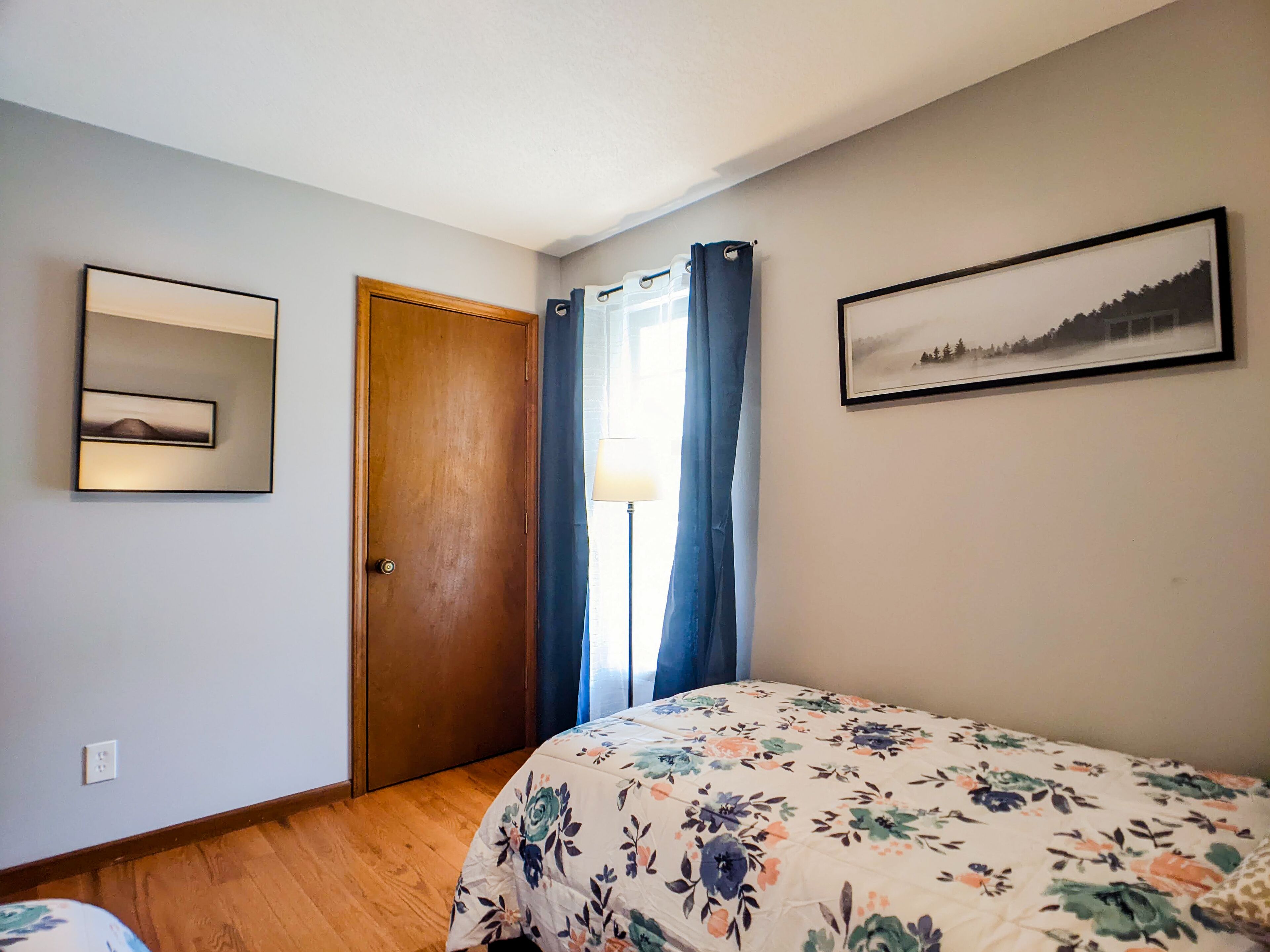 A bedroom with a wooden door, a bed with a floral bedspread, a framed picture on the wall, and a window with blue curtains.