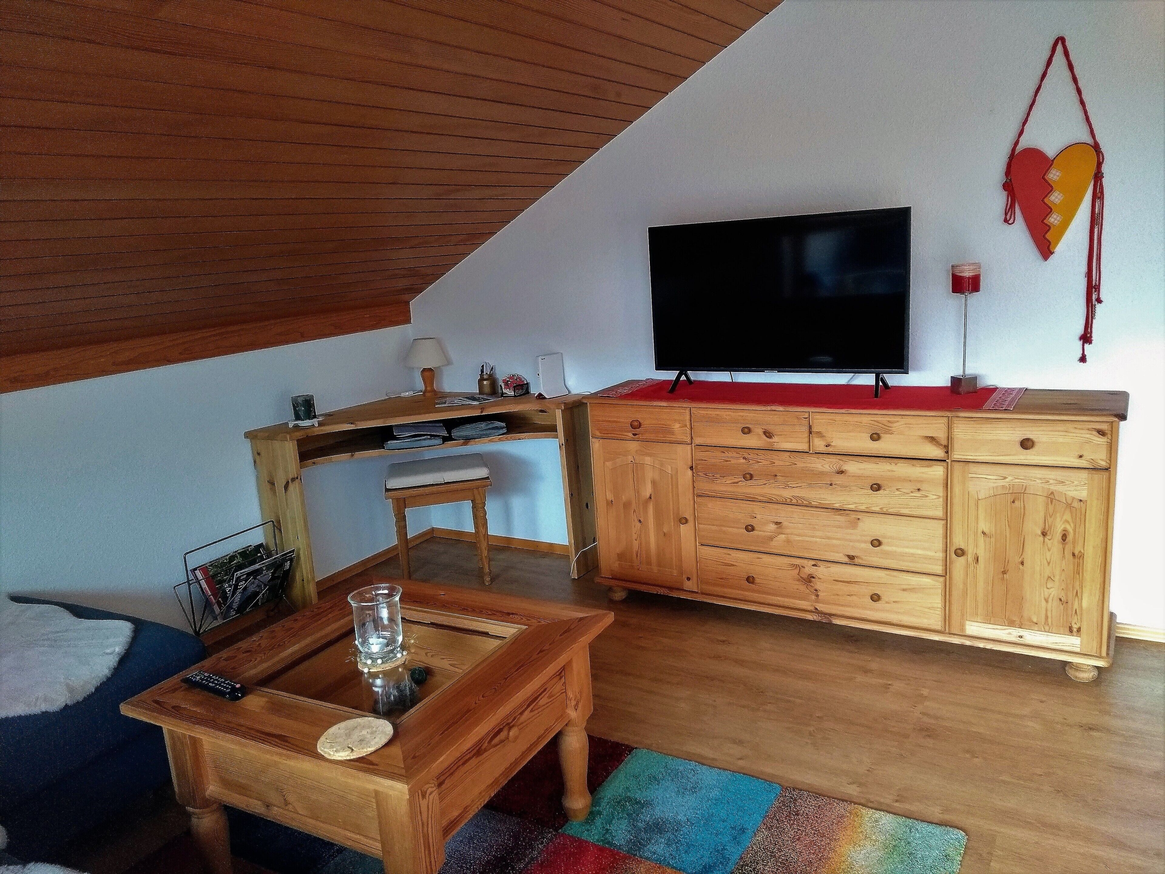 Een gezellige woonruimte met een houten bureau, een aan de muur bevestigde televisie, een klein krukje en een kleurrijk vloerkleed.
