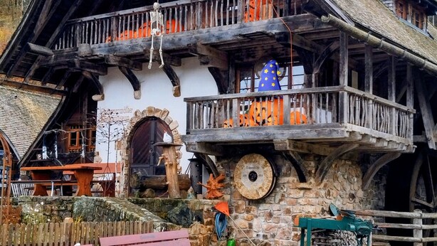 A traditional wooden house with a stone foundation, a balcony with Halloween decorations, and a large wheel visible in the background.