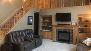Een ruime woonkamer met een open haard, een grote flatscreen-tv, een leren bank en een houten trap.