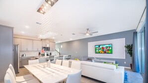 A modern kitchen with white cabinetry, a dining area with a white table and chairs, and a living area with a white sofa and a TV mounted on the wall. - AL1802 Solara Resort 5 Beds 4.5 Baths Private Pool & Free Waterpark (Kissimmee)