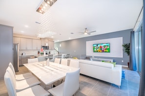 Une cuisine moderne avec des meubles blancs, un coin repas avec une table et des chaises blanches, et un salon avec un canapé blanc et un téléviseur fixé au mur.