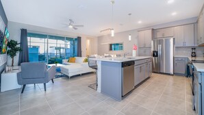 A modern kitchen with a central island, stainless steel appliances, and a dining area with a white sofa and grey armchair. - AL1802 Solara Resort 5 Beds 4.5 Baths Private Pool & Free Waterpark (Kissimmee)