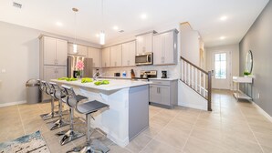 A modern kitchen with a central island, bar stools, stainless steel appliances, and a staircase leading to another level. - AL1802 Solara Resort 5 Beds 4.5 Baths Private Pool & Free Waterpark (Kissimmee)