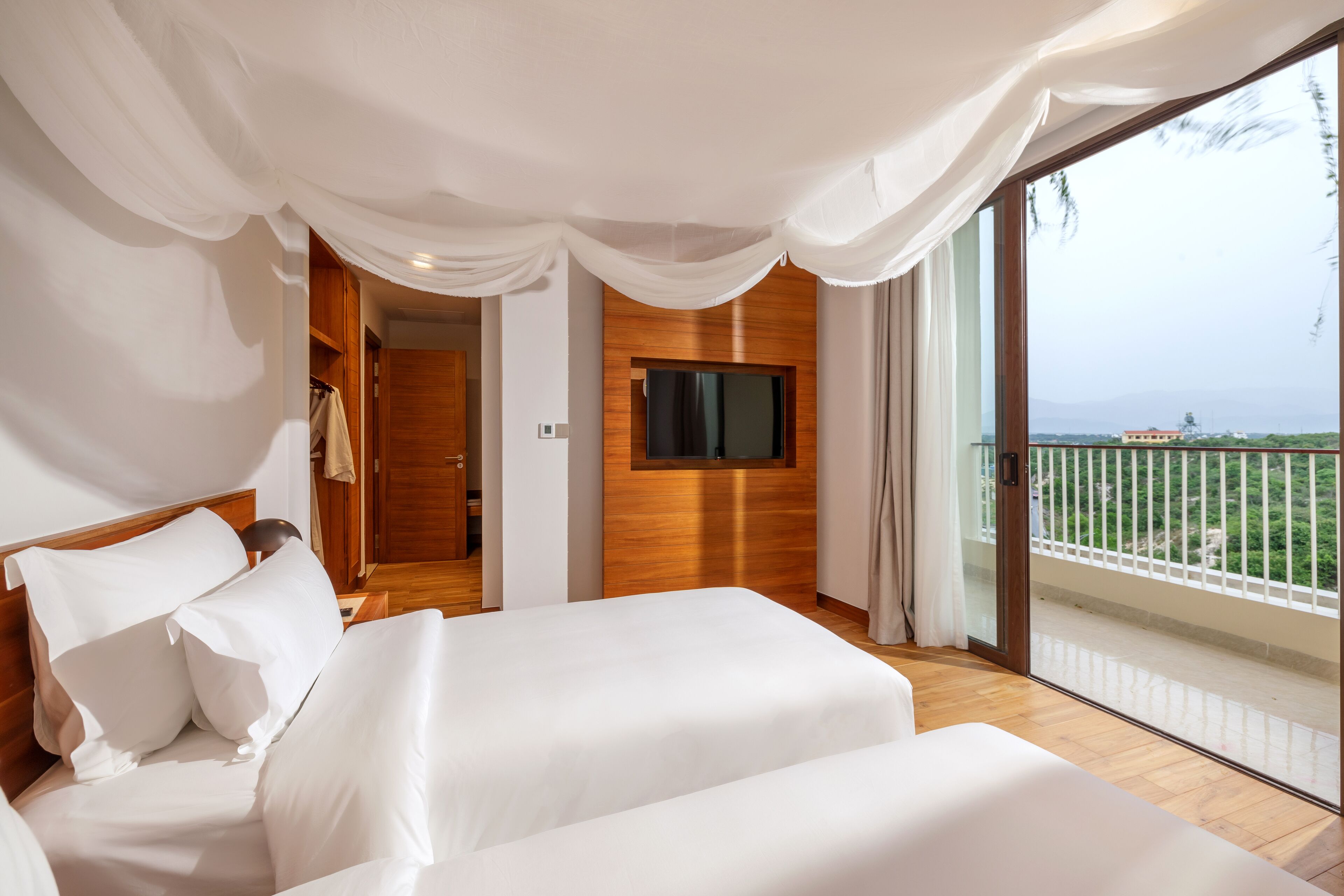 A bedroom with a large bed, a TV mounted on the wall, and a balcony with a view.