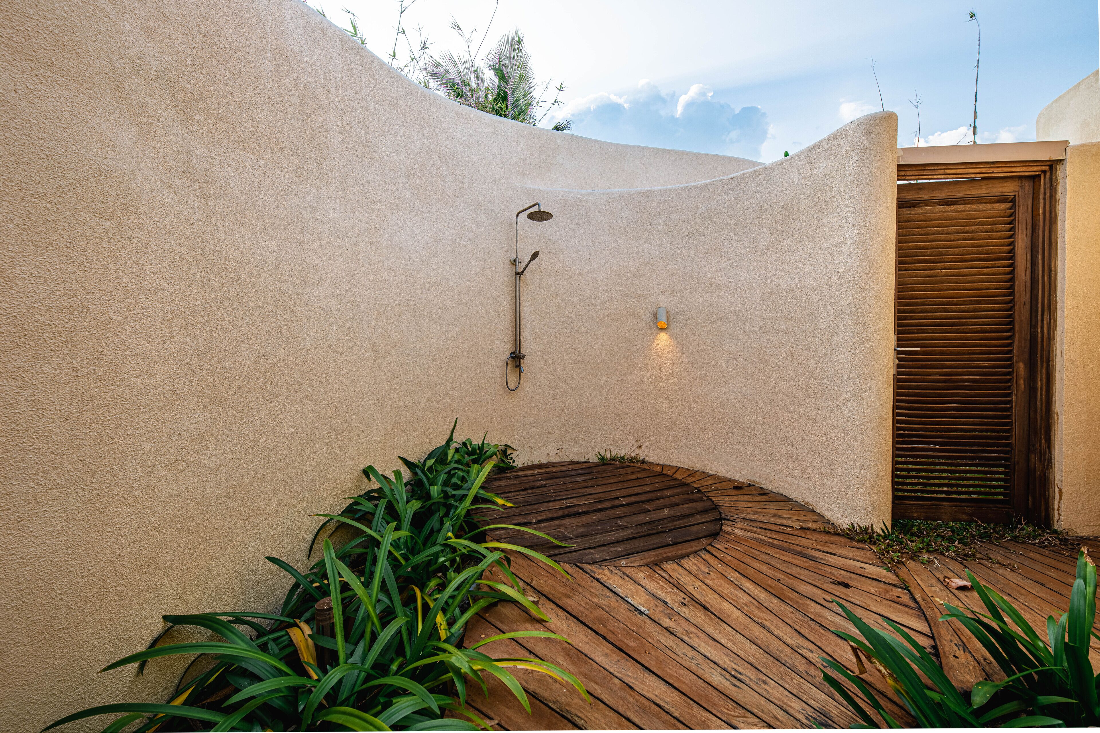 A curved wooden deck with a built-in shower area, surrounded by plants and a beige wall.
