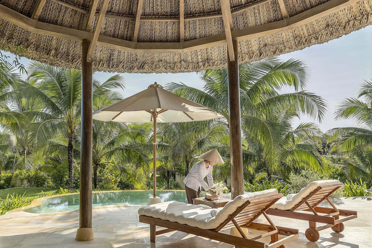 A covered outdoor area with lounge chairs and a pool, surrounded by palm trees.