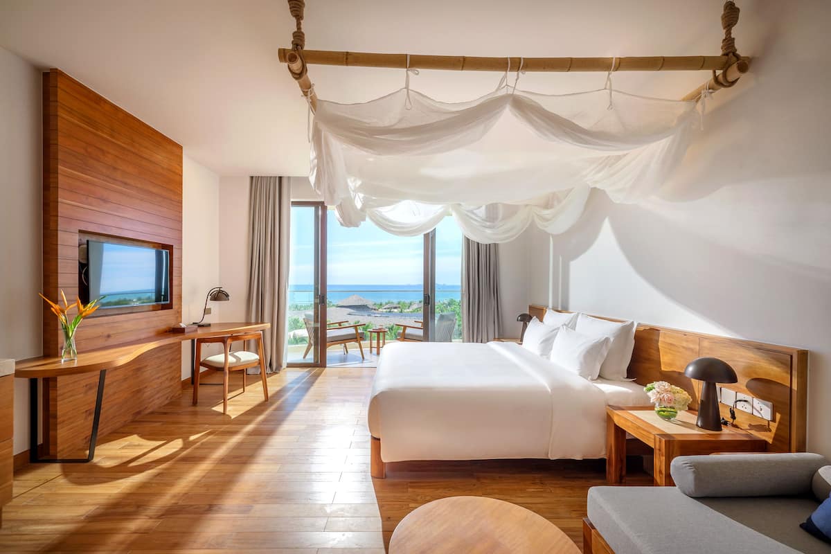 A modern hotel room with a large bed, a wooden desk, a TV, and a view of the ocean.