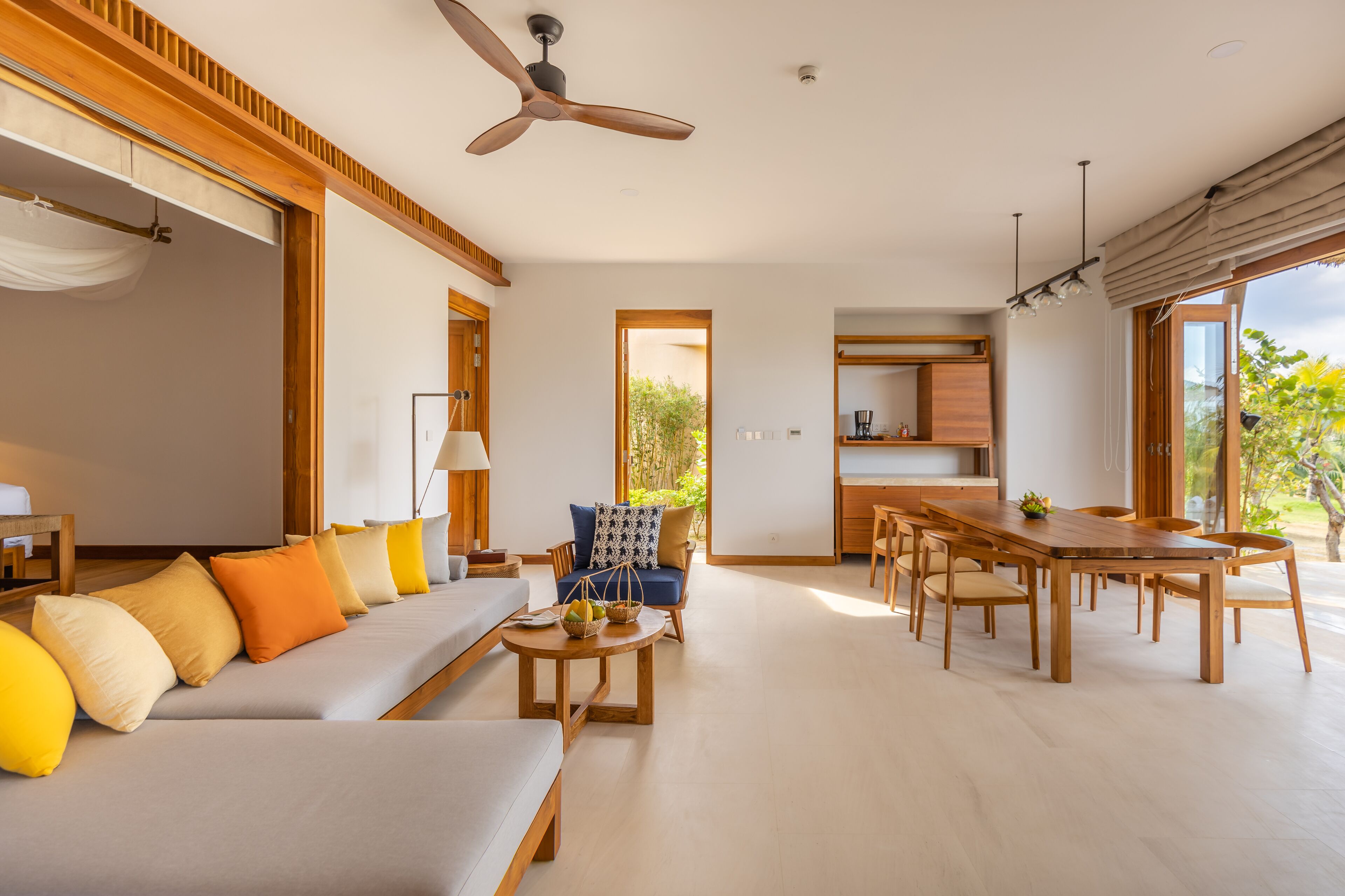 A spacious living area with a sofa, dining table, and chairs, a ceiling fan, and a view of greenery outside.