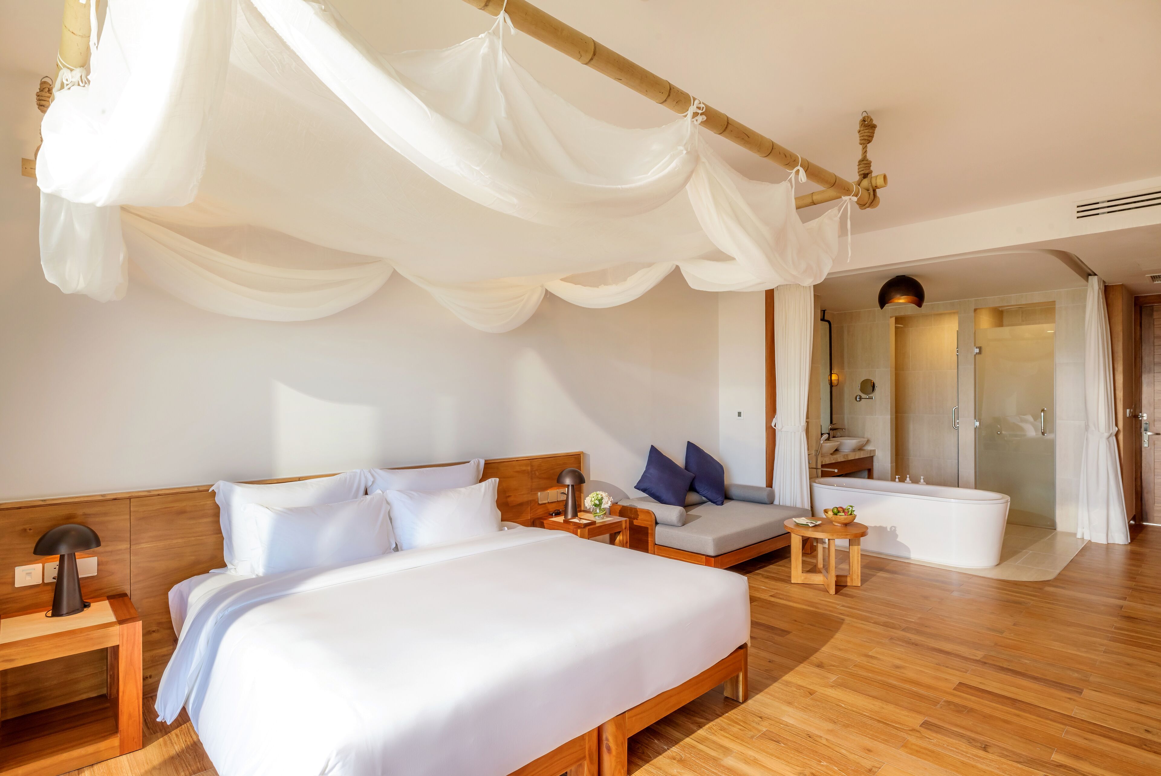 A hotel room with a large bed, a sofa, a small table, a nightstand, a lamp, a bathroom with a bathtub, and a shower.