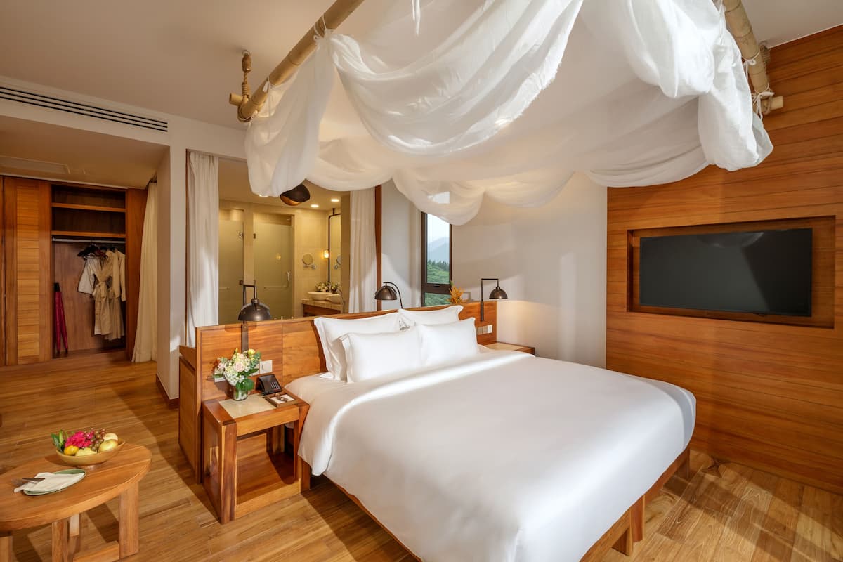 A spacious bedroom with a large bed, wooden headboard, bedside tables with lamps, a wooden wardrobe, and a large window.