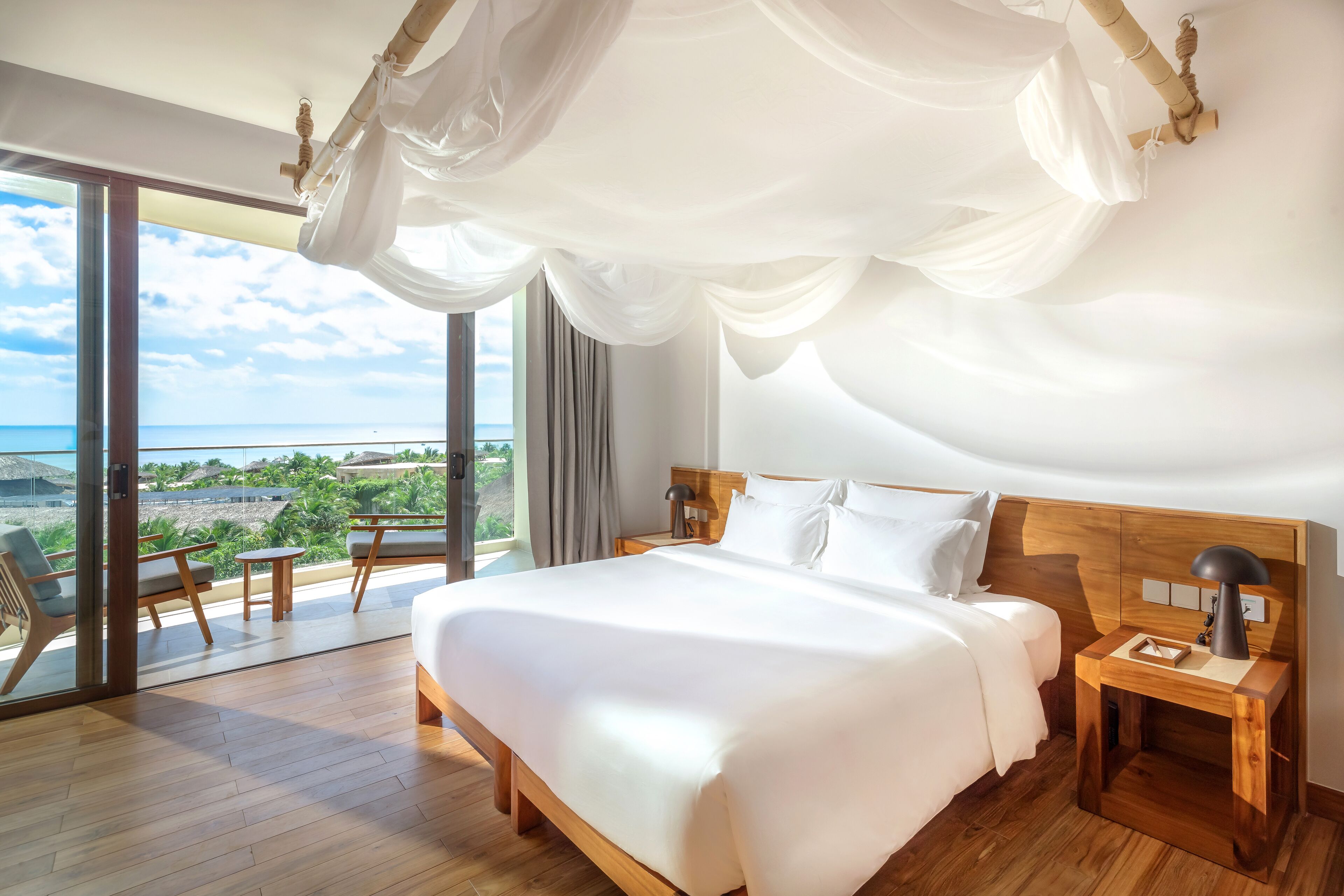 Ein geräumiges Schlafzimmer mit einem großen Bett, einem Holz-Kopfschild und einem Nachttisch mit Lampe. Ein Balkon mit Meerblick ist sichtbar.