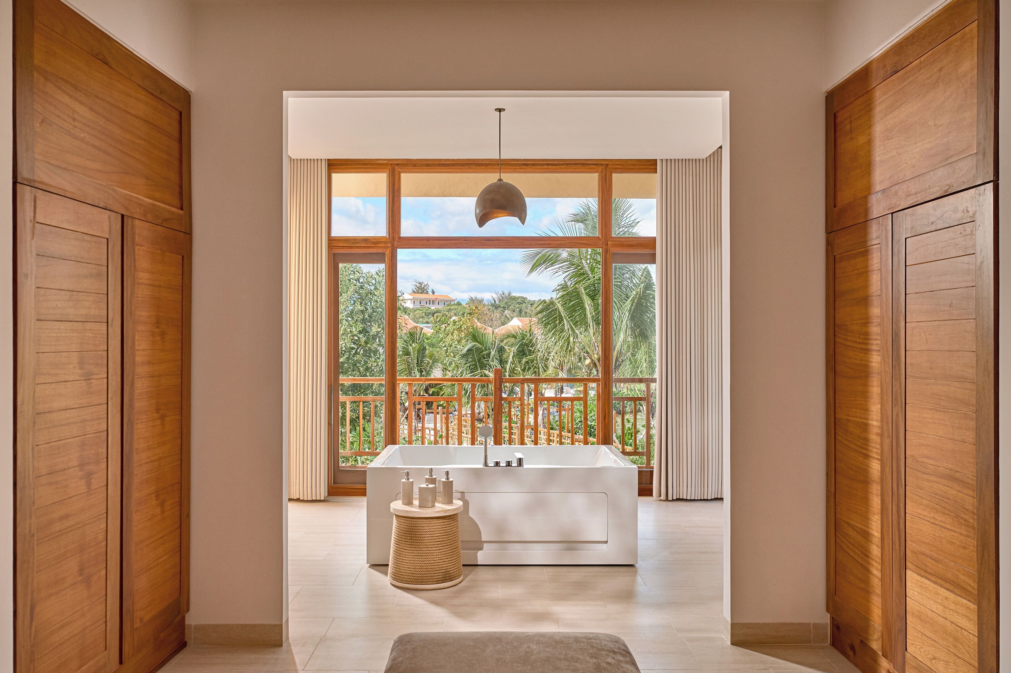 A modern bathroom with a large bathtub, wooden doors, and a view of a tropical landscape.