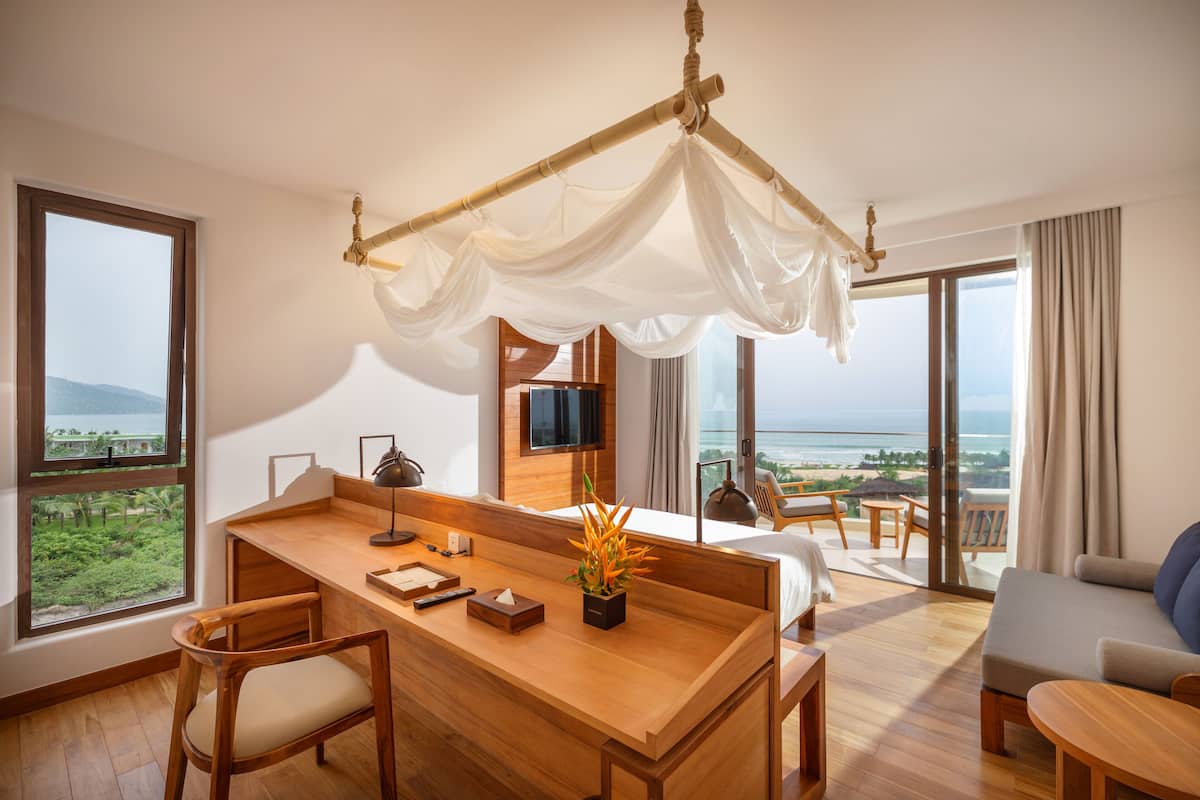 A modern hotel room with a wooden desk, a comfortable sofa, a bed with a canopy, a TV mounted on the wall, and a balcony with a view of the sea.