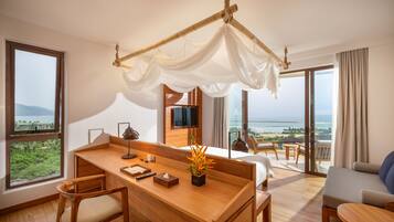 Une chambre d’hôtel moderne avec un bureau en bois, un canapé confortable, un lit à baldaquin, un téléviseur fixé au mur et un balcon avec vue sur la mer.