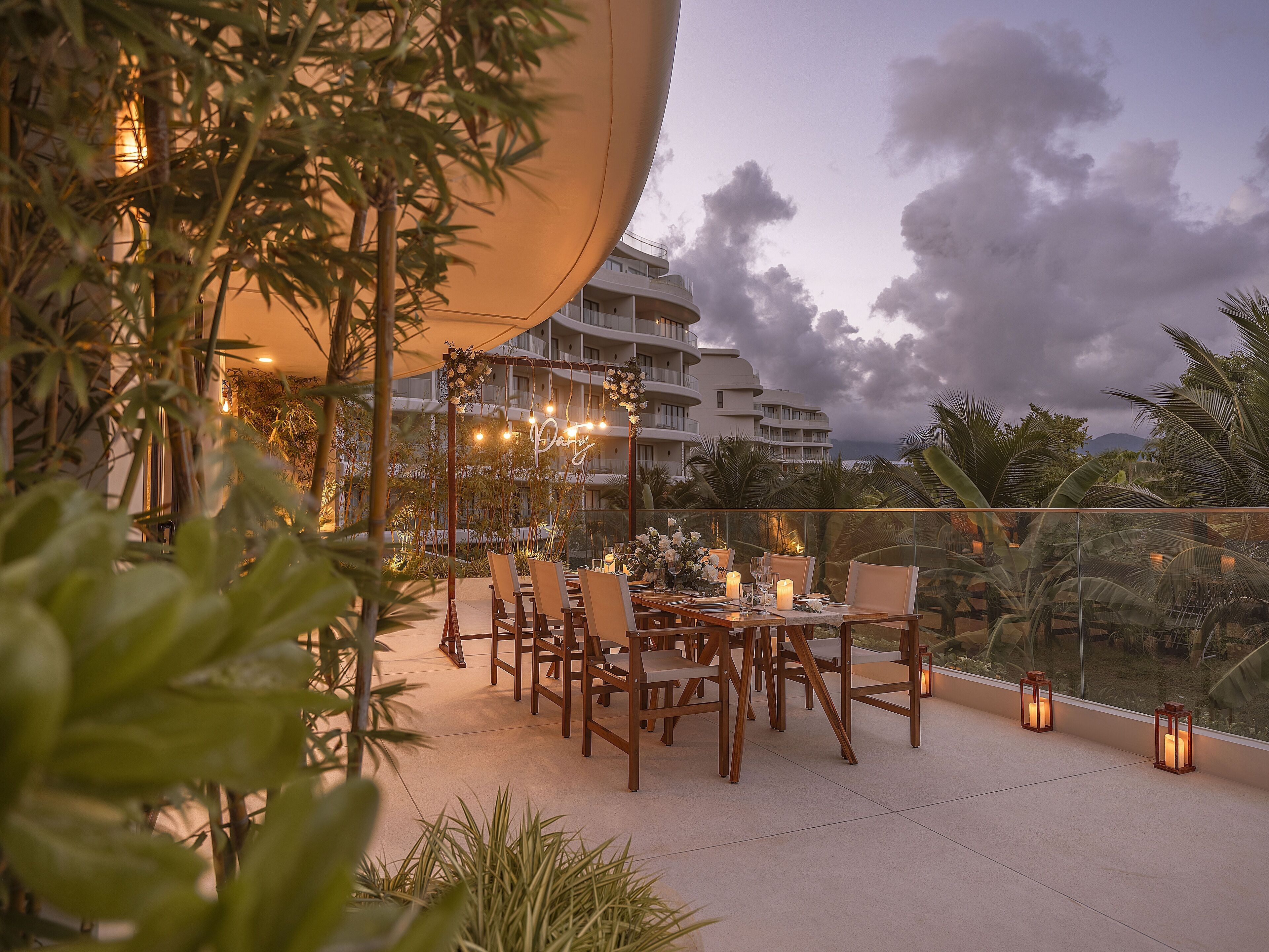 An outdoor dining area with tables and chairs, surrounded by plants and a view of a building with balconies.