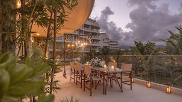 An outdoor dining area with tables and chairs, surrounded by plants and a view of a building with balconies.