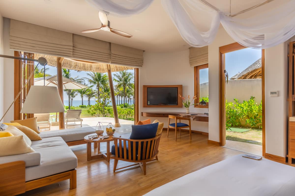 A spacious room with a large bed, a TV, and a desk. There is a balcony with a view of palm trees and the ocean.