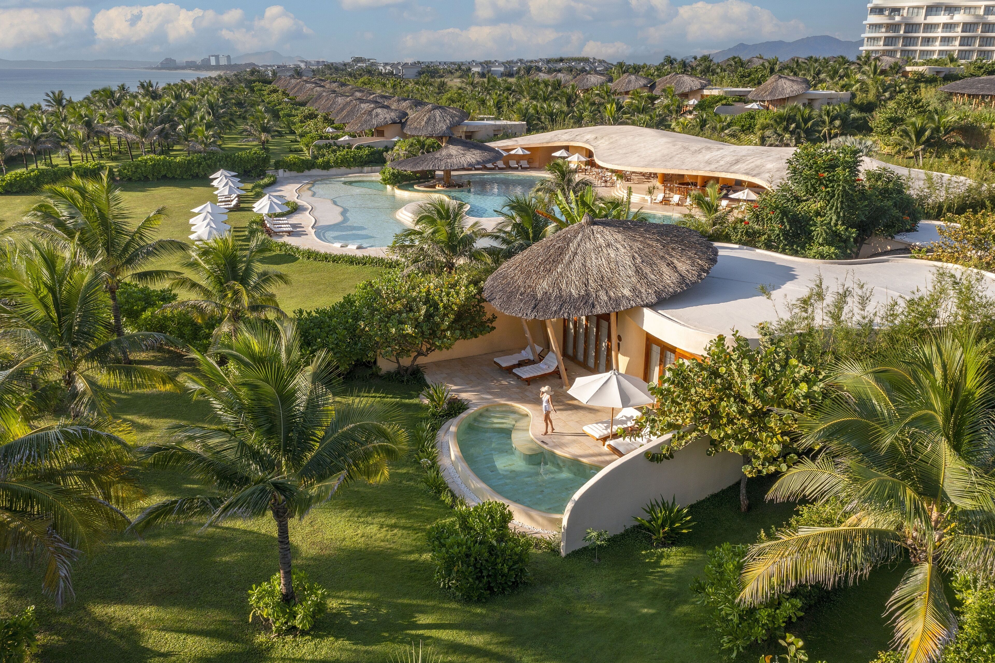 Resort com várias piscinas, palmeiras e construções com telhado de palha.