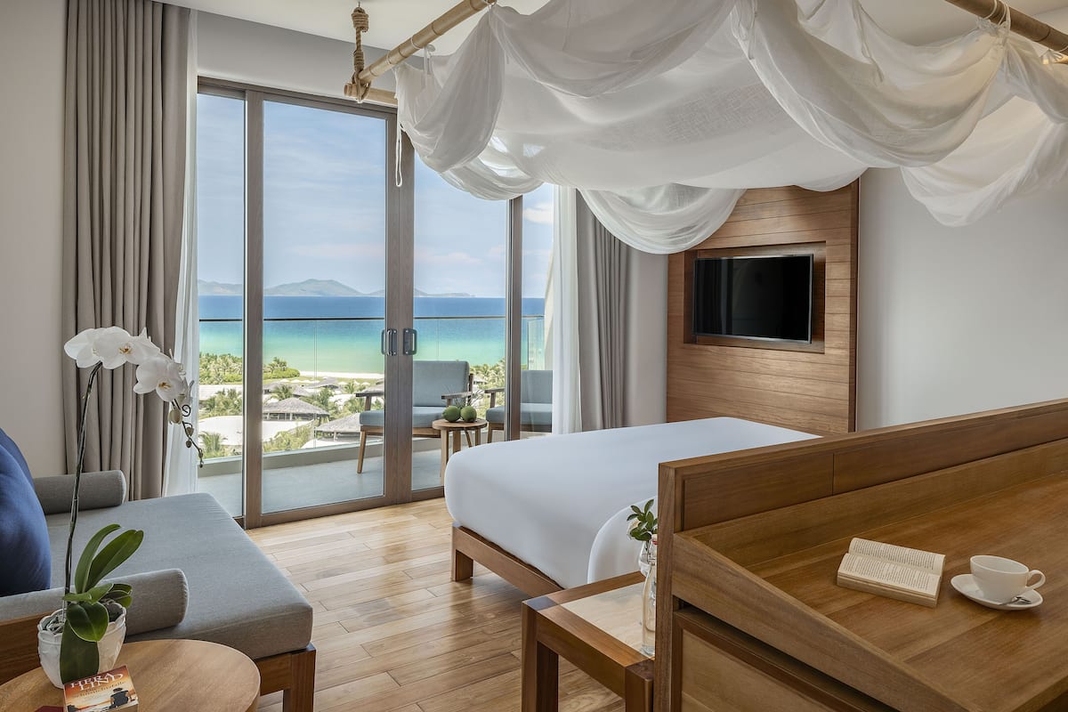 A hotel room with a bed, a TV, a balcony with a view of the sea, and a dining area with a table and chairs.