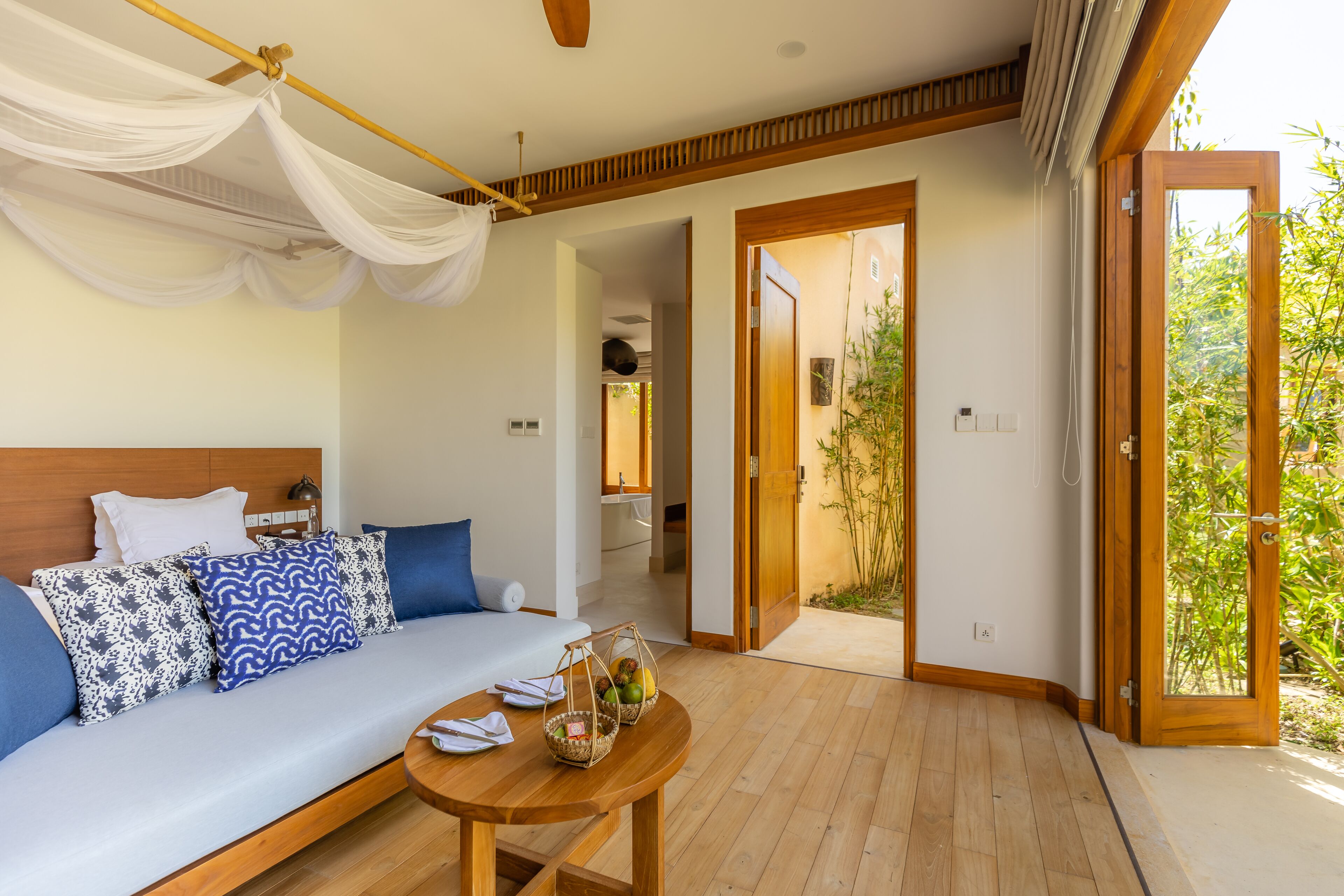 A room with a bed, a table with fruit baskets, a wooden floor, and a door leading to a garden.