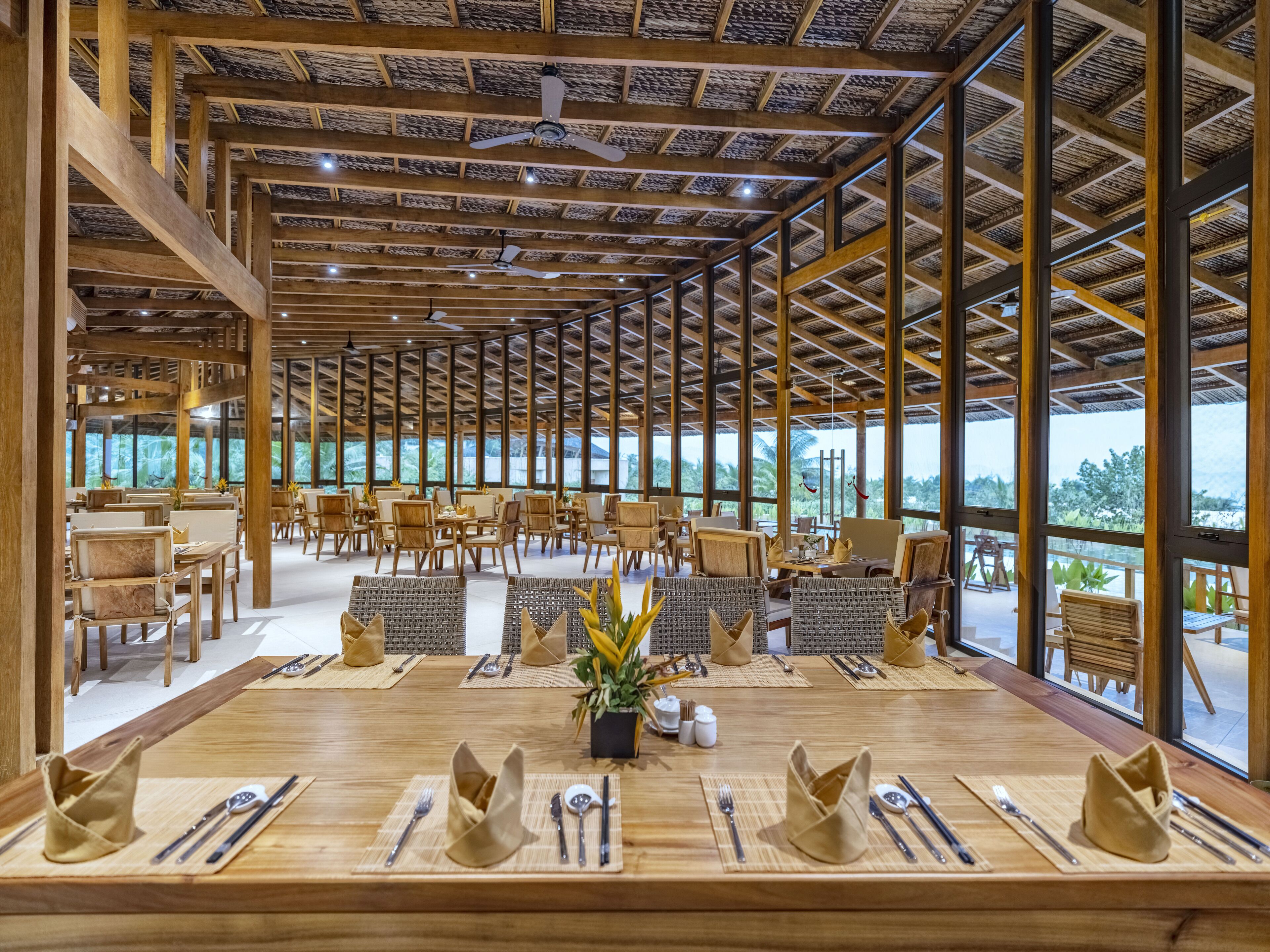 A spacious dining area with wooden tables and chairs, large windows offering a view of the outdoors, and a high ceiling with exposed beams.
