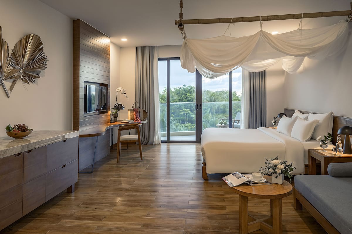 A modern hotel room with a large bed, a desk, a seating area, and a balcony with a view.