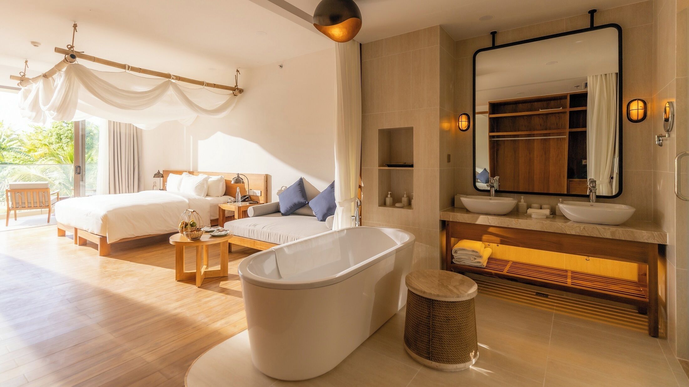A modern hotel room with a large bed, a sitting area, a bathtub, and a bathroom with a large mirror and sink.