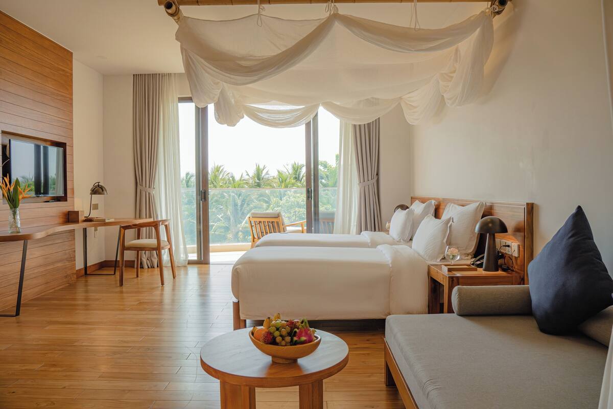 A hotel room with two beds, a desk, a chair, a round table with a fruit bowl, and a balcony with curtains.