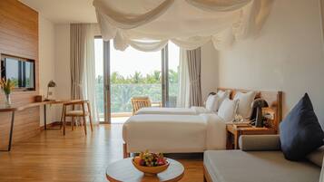 A hotel room with two beds, a desk, a chair, a round table with a fruit bowl, and a balcony with curtains.