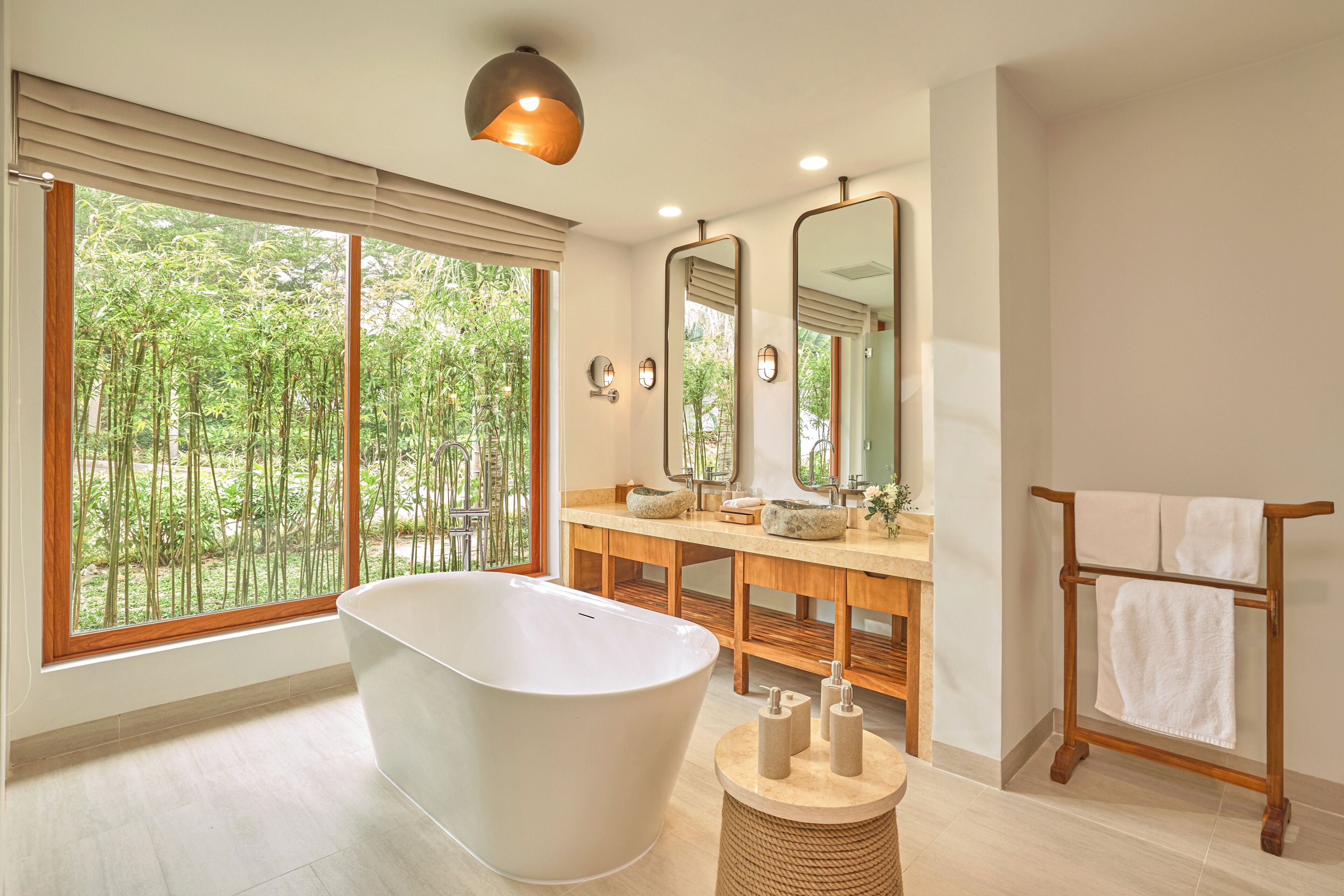 Une salle de bain moderne avec une baignoire sur pieds, un meuble-lavabo en bois, de grands miroirs et une vue sur la verdure grâce à de grandes fenêtres.