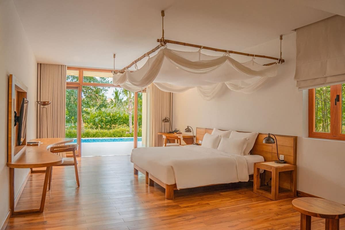 A bedroom with a bed, bedside tables, a desk, and a chair. There is a large window with a view of greenery.